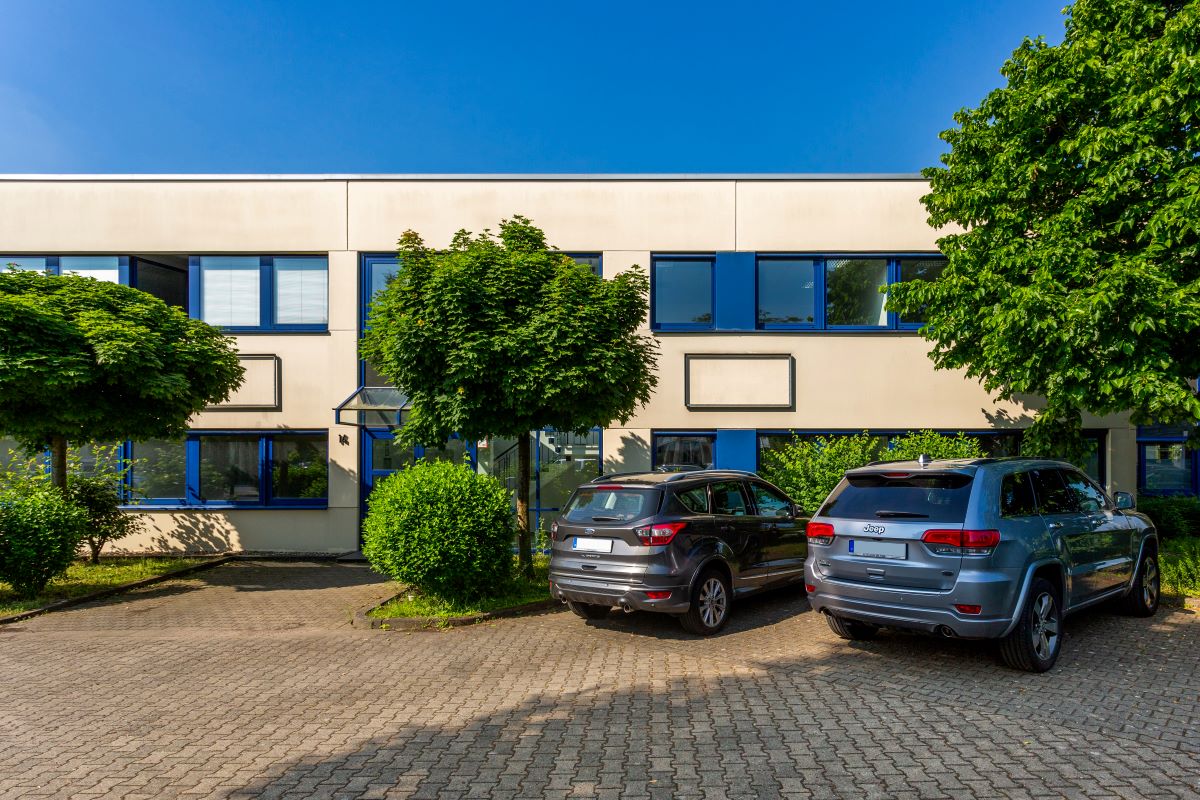 Ein modernes Bürogebäude mit blau umrandeten Fenstern, Bäumen, Büschen und zwei geparkten Geländewagen im Vordergrund unter einem klaren blauen Himmel.