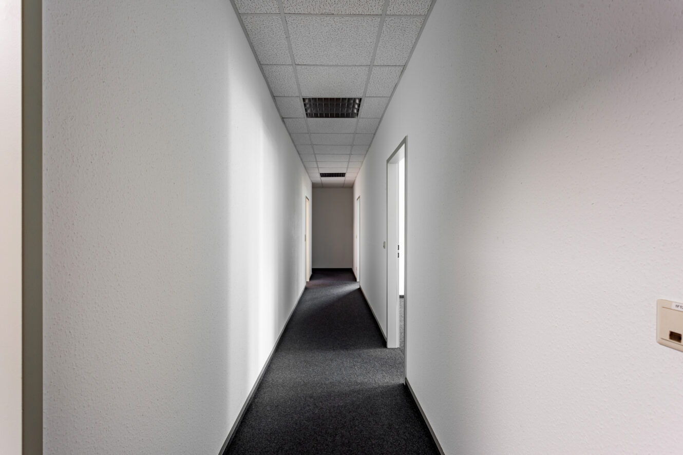 Ein langer, leerer Flur mit weißen Wänden, dunklem Teppichboden und Neonröhren an der Decke; die Türen sind auf beiden Seiten offen.