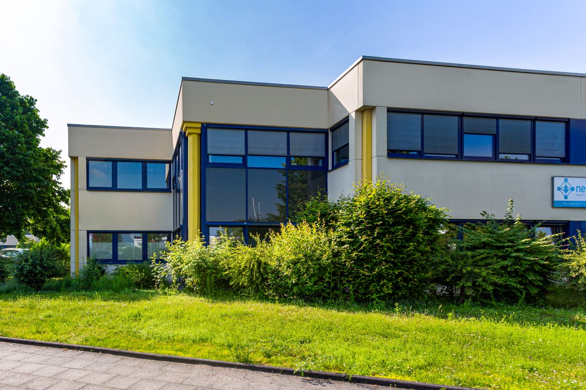 Ein modernes zweistöckiges Bürogebäude mit großen Fenstern, beigen Wänden und gelben Säulen, umgeben von grünem Gras und Büschen an einem sonnigen Tag.