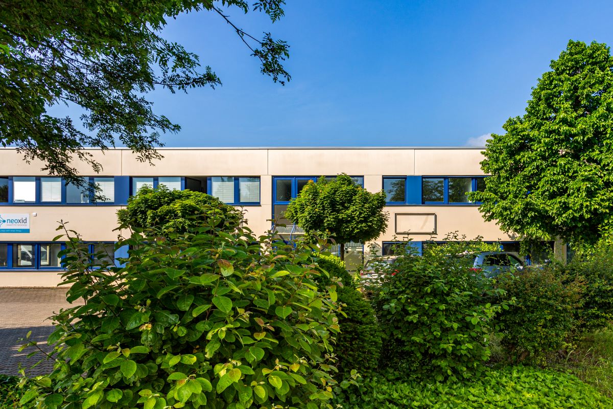 Modernes zweistöckiges Bürogebäude mit blau umrahmten Fenstern, umgeben von grünen Bäumen und Sträuchern unter strahlend blauem Himmel.