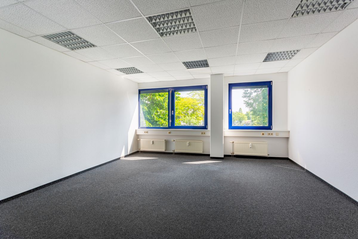 Leerer Büroraum mit weißen Wänden, grauem Teppich und zwei blau umrahmten Fenstern, die natürliches Licht hereinlassen; Heizkörper und Steckdosen unter den Fenstern.
