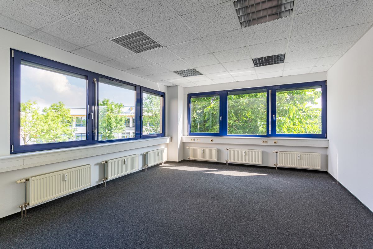 Leerer Büroraum mit großen, blau gestrichenen Fenstern, weißen Wänden, grauem Teppich und Heizkörpern unter den Fenstern. Helles Tageslicht erhellt den Raum.