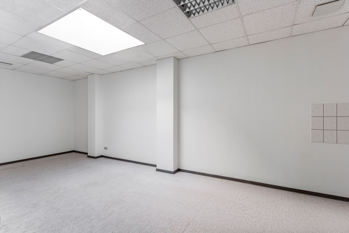 Leerer Raum mit weißen Wänden, weißen Deckenfliesen und grauem Bodenbelag; mit Deckenbeleuchtung und einem kleinen gefliesten Bereich an einer Wand.