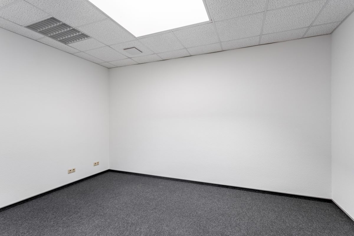 Leerer Raum mit weißen Wänden, grauem Teppich und einer weiß gekachelten Decke mit Leuchtstoffröhren. An der linken Wand sind zwei Steckdosen zu sehen.