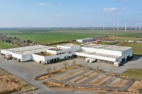 Luftaufnahme eines großen, weißen industriellen Lagerkomplexes, umgeben von leeren Parkplätzen, Feldern und Windrädern im Hintergrund.