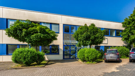 Ein modernes zweistöckiges Bürogebäude mit großen blauen Fenstern, umgeben von Büschen und Bäumen und zwei geparkten Autos davor.