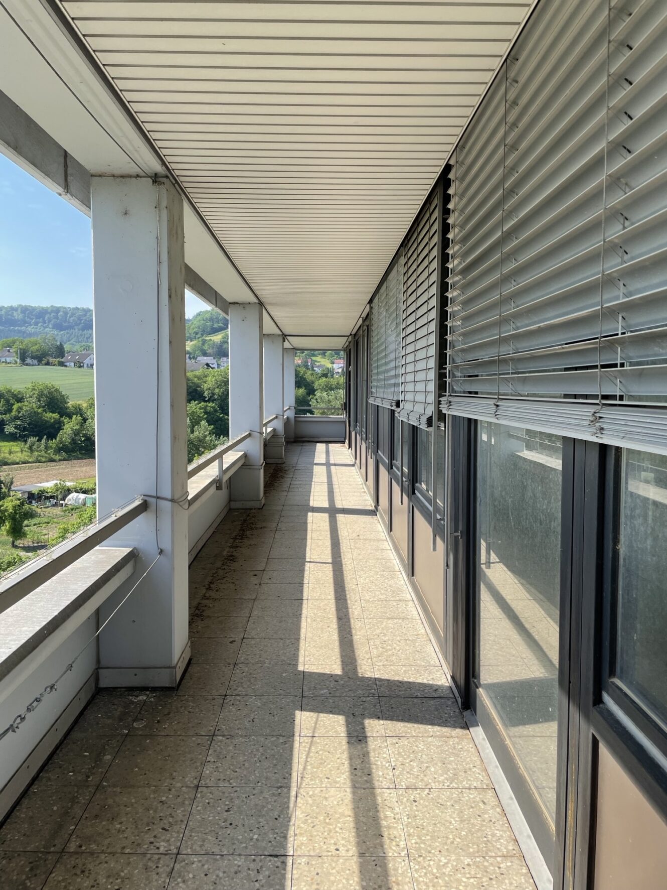 Ein langer Außenbalkon mit gefliestem Boden, Metallgeländer, Säulen und großen Fenstern mit geschlossenen Jalousien an einem sonnigen Tag. Im Hintergrund ist eine grüne Landschaft zu sehen.