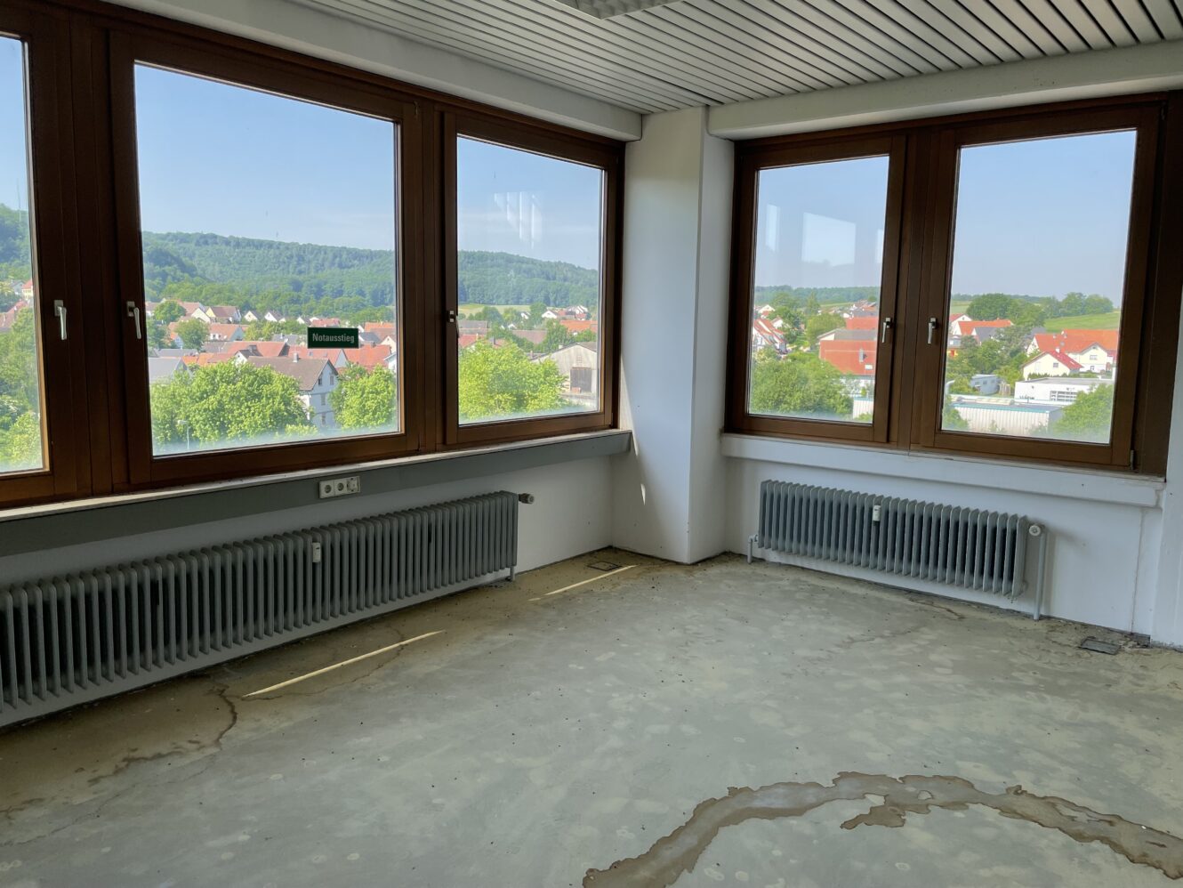 Ein leerer Raum mit großen Fenstern, Sichtbetonboden und zwei Heizkörpern. Aus den Fenstern blickt man auf eine Stadt mit Bäumen und Hügeln im Hintergrund.