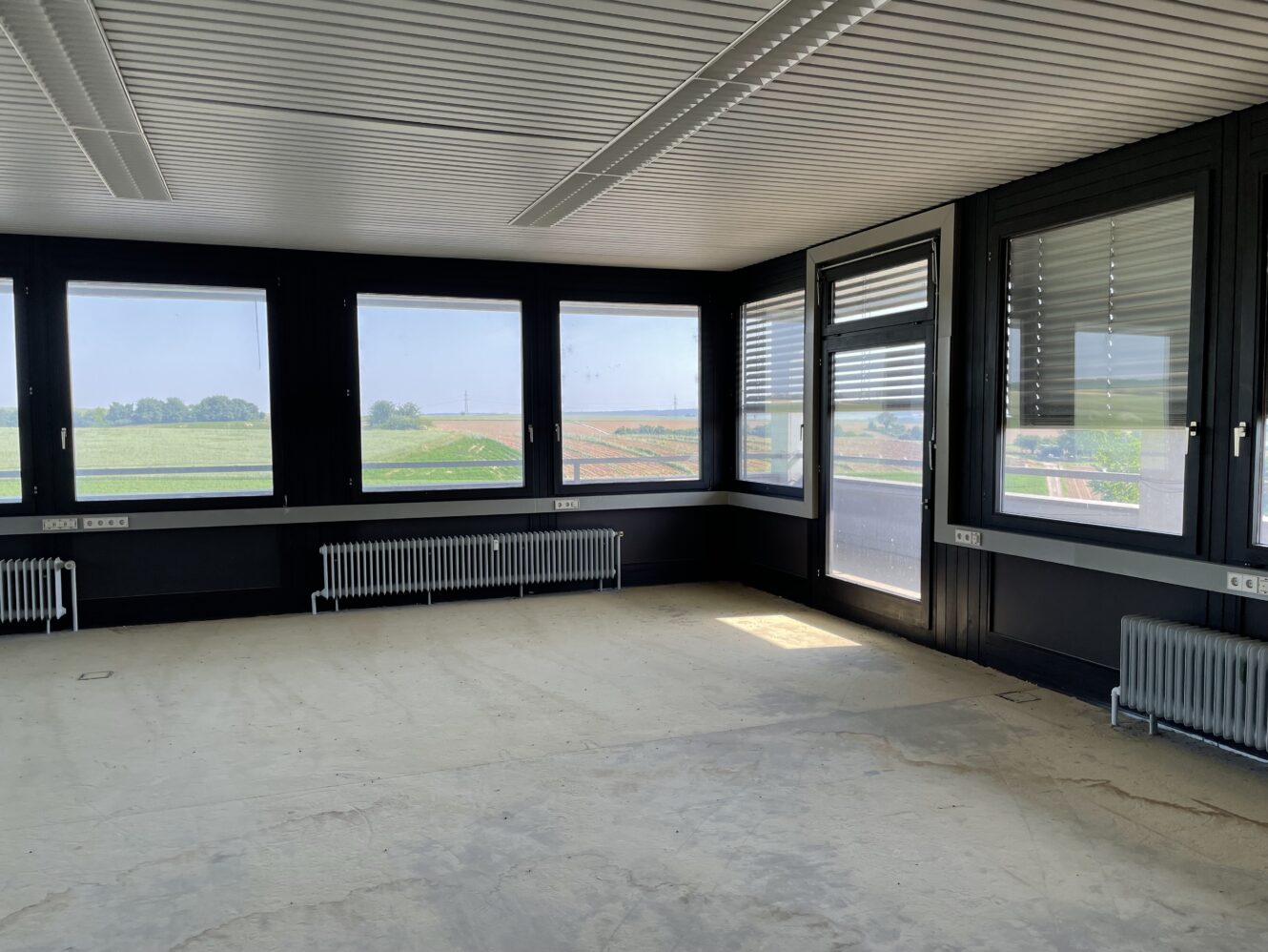 Leerer Büroraum mit großen Fenstern, schwarzen Rahmen, Heizkörpern unter den Fenstern und einem nackten Fußboden; durch das Glas ist die Außenlandschaft sichtbar.