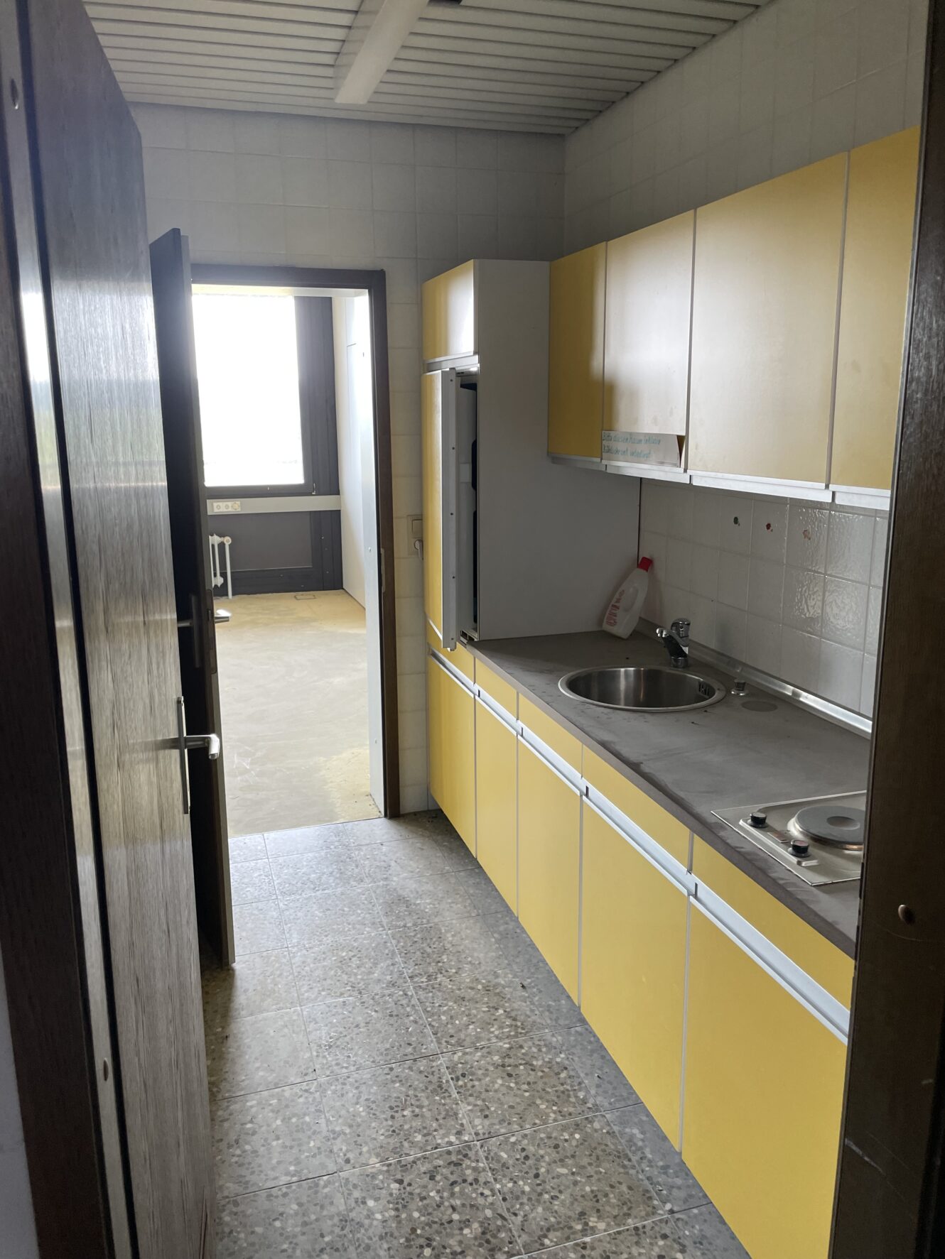 Schmale Küche mit gelben Schränken, Edelstahlspüle, Elektroherd und Putzmitteln auf dem Tresen; eine offene Tür führt in einen helleren Nebenraum.