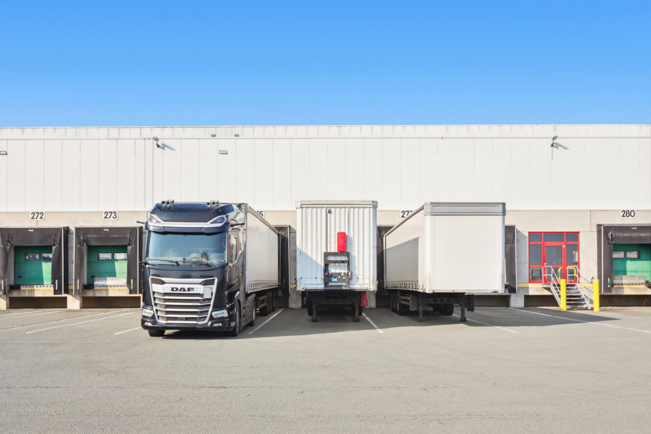 An einem klaren Tag sind drei große Lastwagen an den Laderampen vor einem weißen Industrielager geparkt.