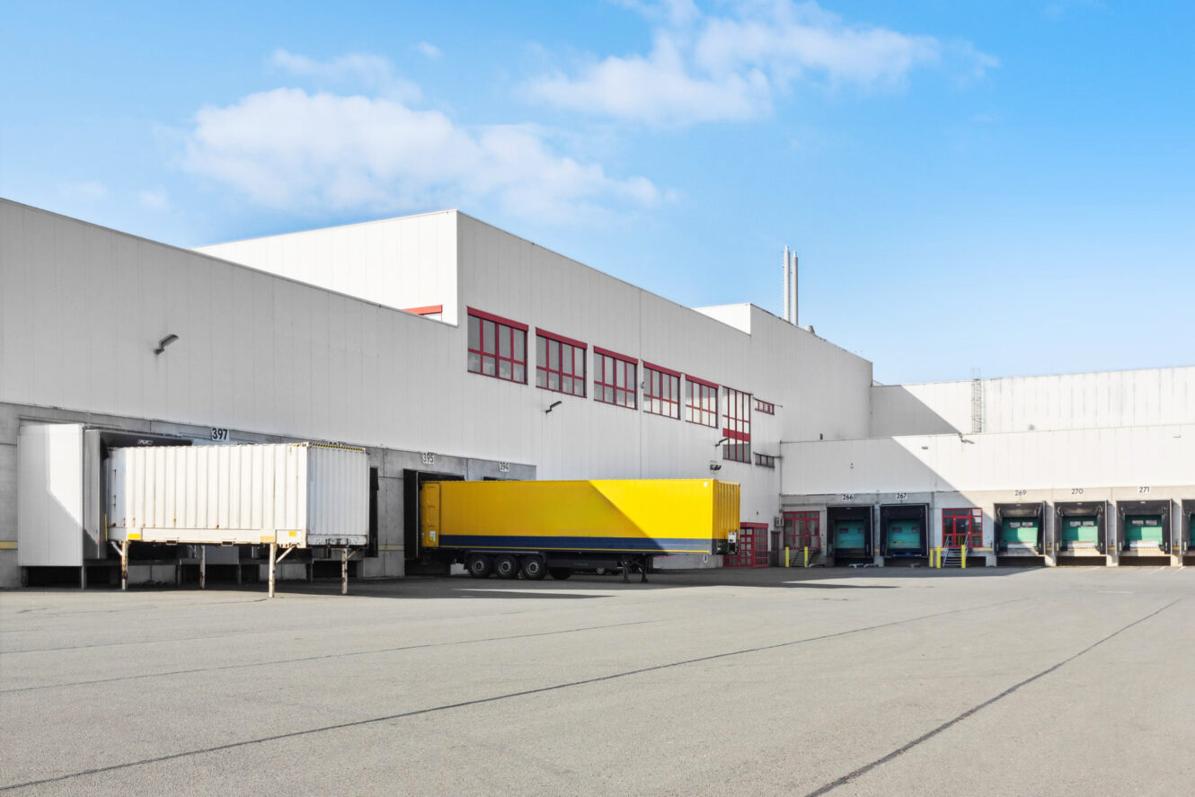 Ein großes Lagerhaus mit mehreren Laderampen; ein gelber Sattelauflieger und ein weißer Anhänger sind unter strahlend blauem Himmel an den Rampen geparkt.