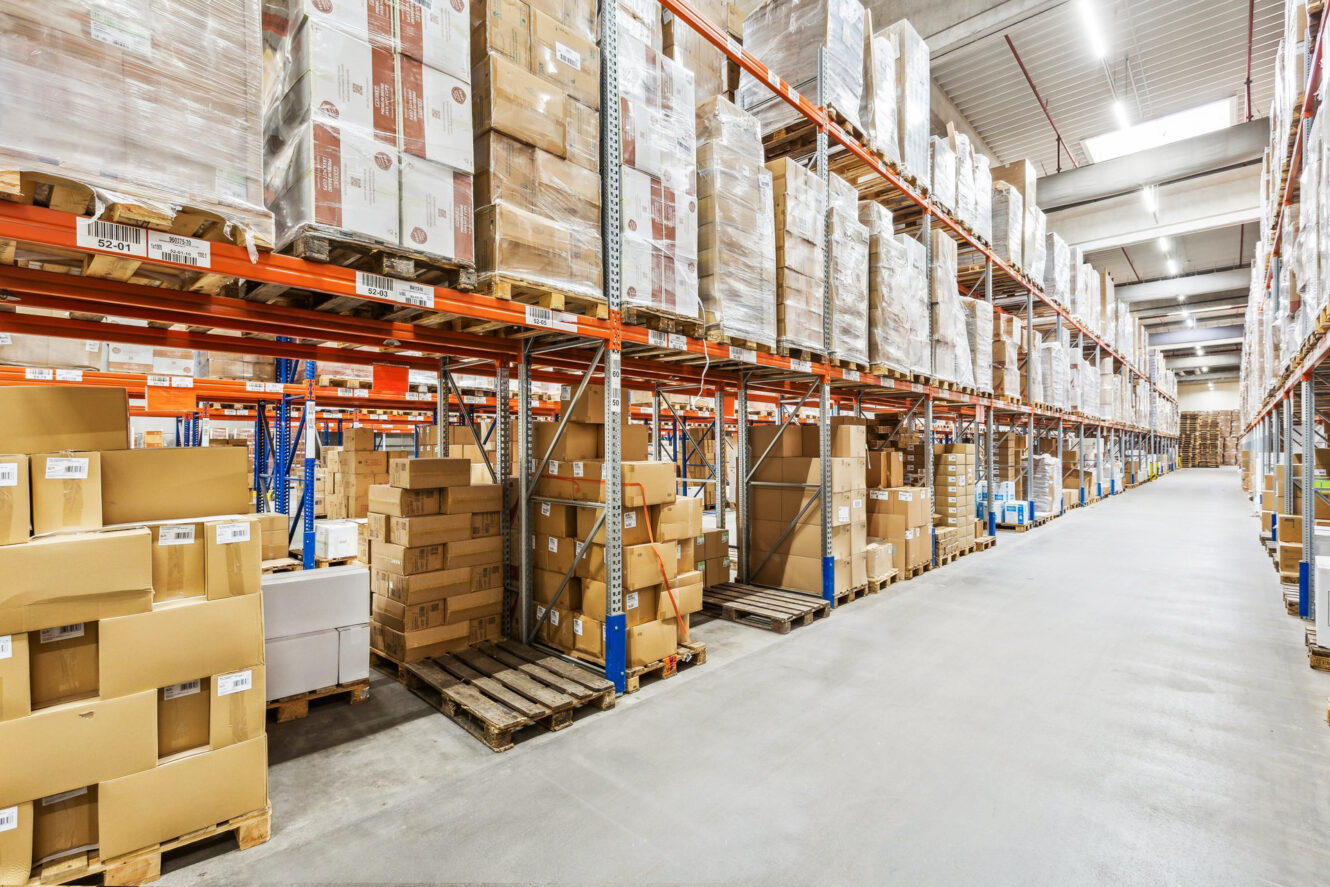 Lagerhalle mit hohen Regalen, in denen Kartons und Paletten gestapelt sind, um geordnete Gänge für die Lagerung und Verteilung zu schaffen.