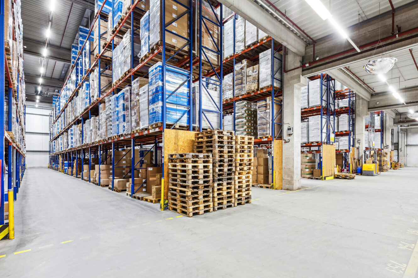Ein großes Lagerhaus mit hohen Regalen mit Kisten, in der Mitte gestapelten Paletten und breiten Gängen, in denen man sich bewegen kann.