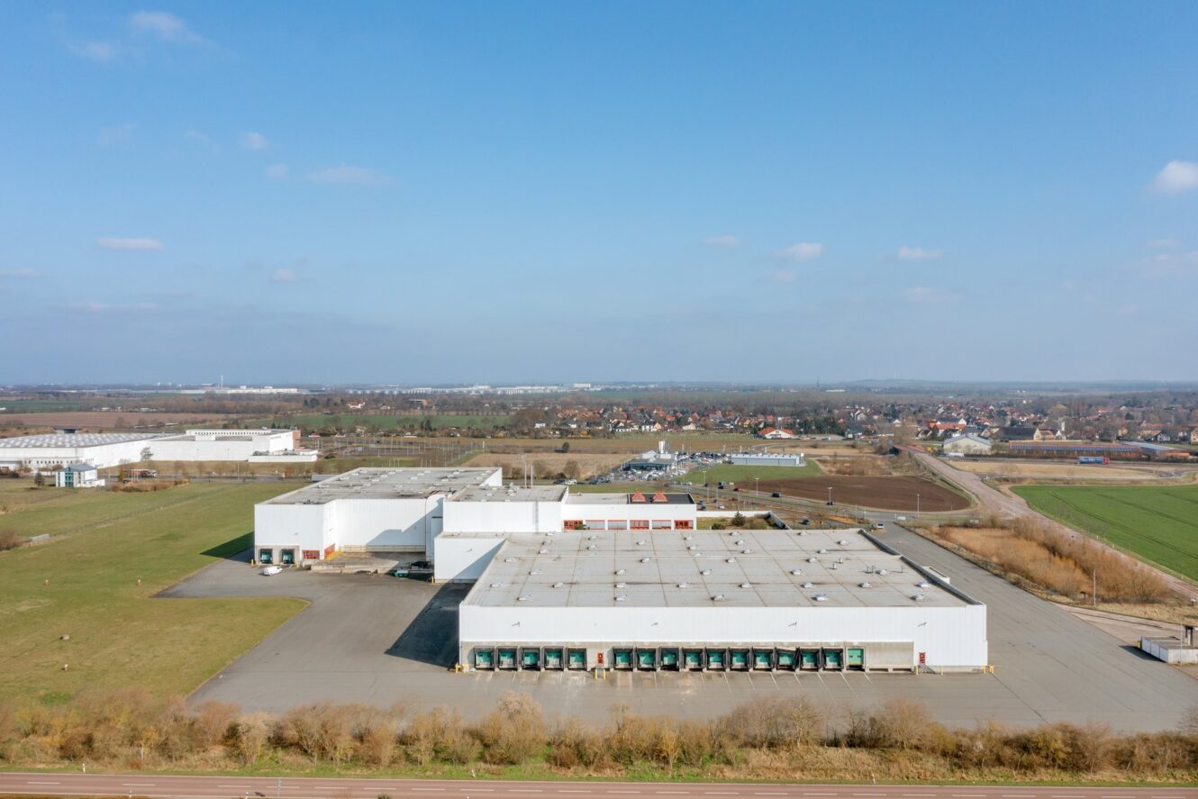 Luftaufnahme eines großen industriellen Lagerkomplexes mit mehreren weißen Gebäuden, Laderampen und umliegenden Freiflächen an einem klaren Tag.