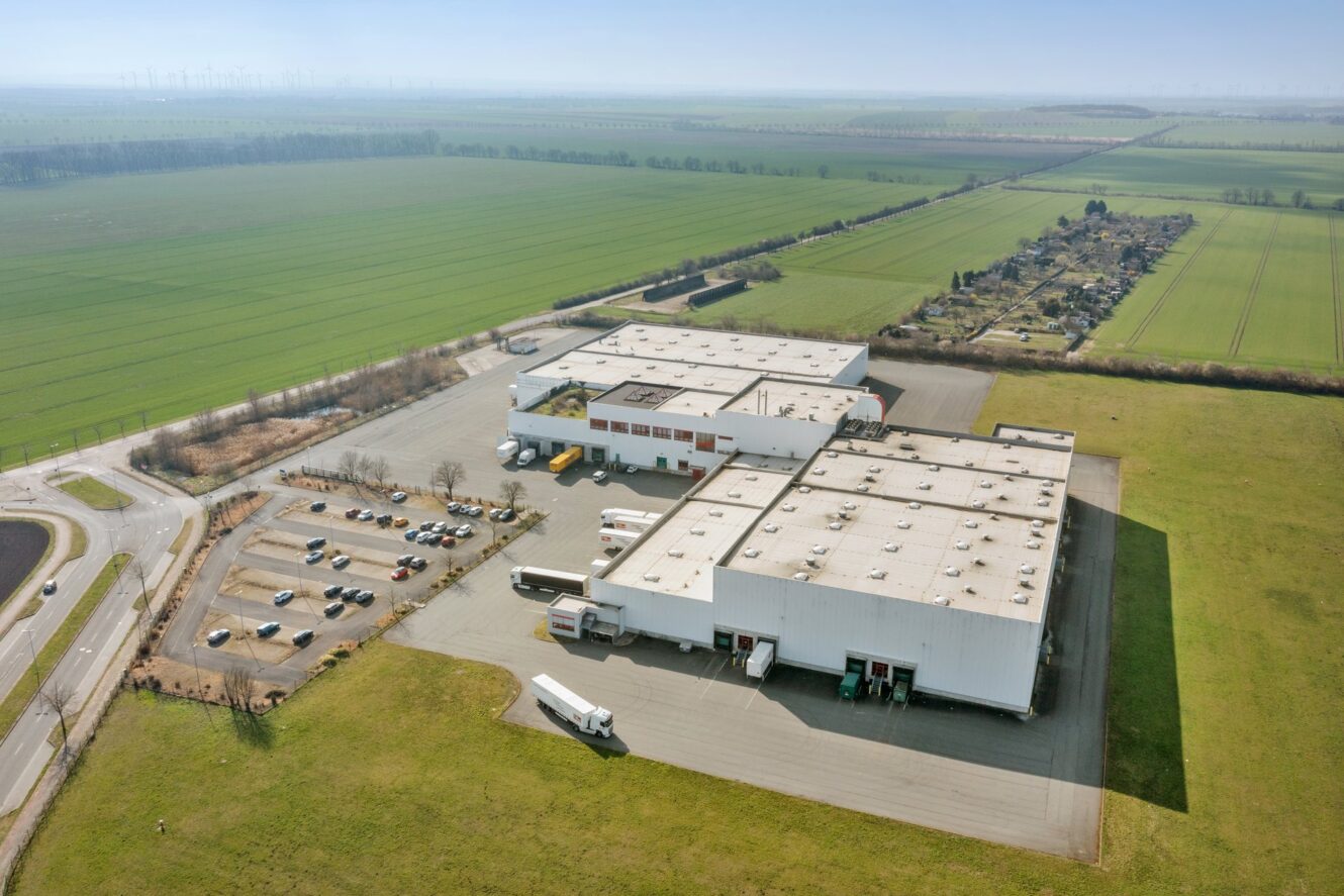 Luftaufnahme eines großen industriellen Lagerkomplexes mit Lastwagen, geparkten Autos und umliegenden grünen Feldern in einer ländlichen Gegend.