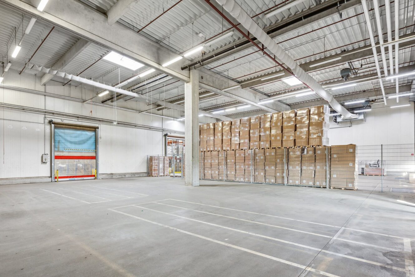 Ein geräumiges Lagerhaus mit gestapelten Paletten von Kartons entlang einer Wand, einer geschlossenen Ladetür und markierten Parkplätzen auf dem Betonboden.