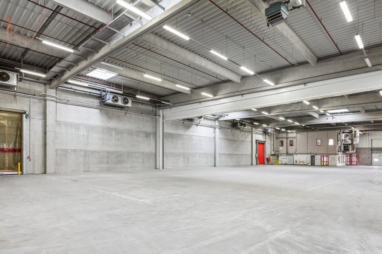 Geräumiges, leeres Industrielager mit Betonböden, hohen Decken, freiliegenden Rohren und großen Oberlichtern. Rote Türen und Fenster säumen die Rückwand.