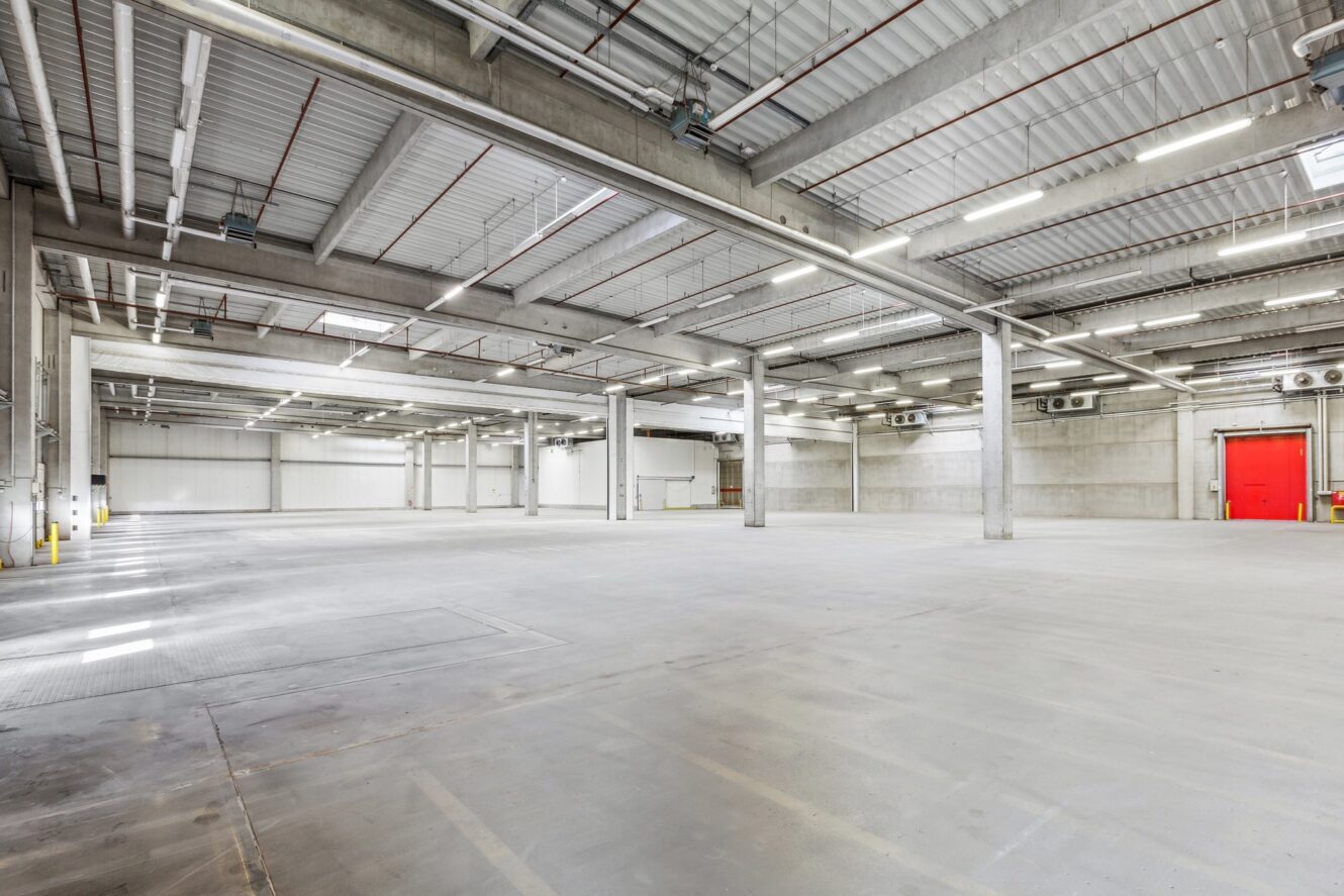 Ein geräumiges, leeres Industrielager mit Betonböden, hohen Decken, freiliegenden Balken, Leuchtstoffröhren und einer roten Tür am anderen Ende.