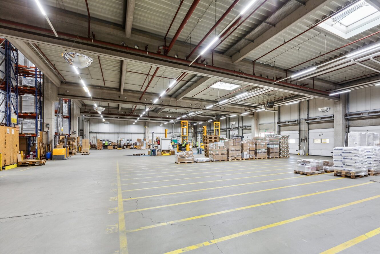 Geräumige Lagerhalle mit hohen Decken, Paletten mit Waren, Industrieregalen und gelben Sicherheitslinien auf dem Boden.
