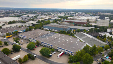 Luftaufnahme eines Industriegebiets mit großen Lagerhallen, geparkten Lastwagen, Bürogebäuden und umliegenden Grünflächen unter einem bewölkten Himmel.