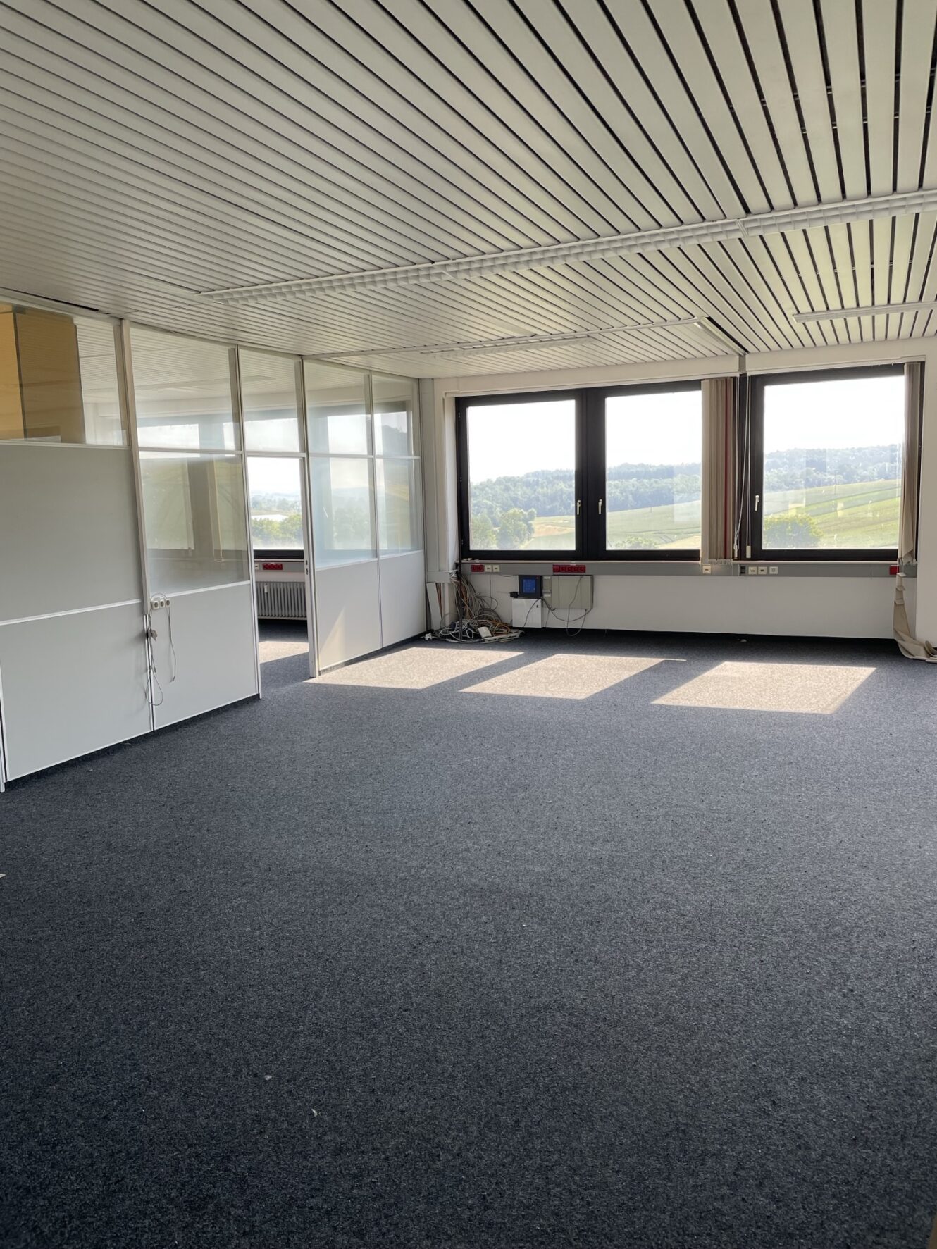 Ein großer, leerer Büroraum mit Teppichboden, weißen Trennwänden auf der linken Seite, Fenstern auf der rechten Seite, die das Sonnenlicht hereinlassen, und ein paar Kabeln und kleinen Gegenständen in der Nähe der Fenster.