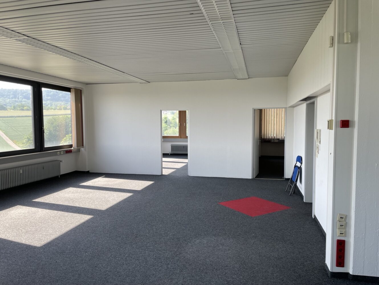 Ein leerer Büroraum mit grauem Teppich, weißen Wänden, großen Fenstern und einem einzigen roten Teppichplatz. Ein blauer Stuhl steht im angrenzenden Raum, der durch eine Türöffnung sichtbar ist.