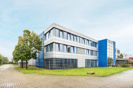 Modernes dreistöckiges Bürogebäude mit blau-weißer Fassade, großen Fenstern und einer kleinen Rasenfläche davor, fotografiert an einem klaren Tag.