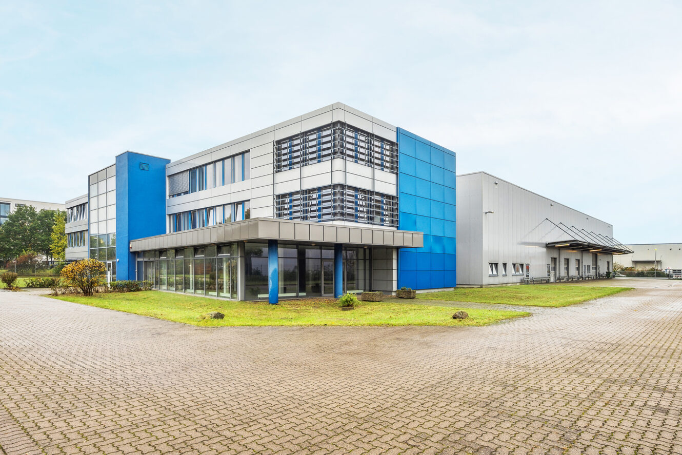 Modernes Industriegebäude mit blauem und grauem Äußeren, großen Fenstern und einer Laderampe, auf einem gepflasterten Grundstück mit etwas Gras und kleinen Bäumen gelegen.