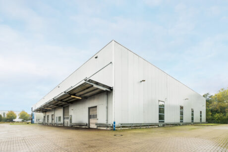 Ein großes, weißes Industrielager mit Ladebuchten und Überdachungen auf einem gepflasterten Grundstück unter einem teilweise bewölkten Himmel.