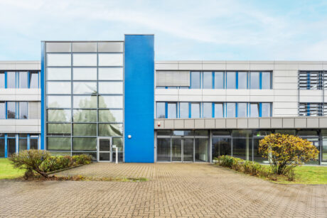 Modernes dreistöckiges Bürogebäude mit einem blauen vertikalen Akzent, großen Glasfenstern und einem gepflasterten Vorhof mit einigen Grünpflanzen und Sträuchern.