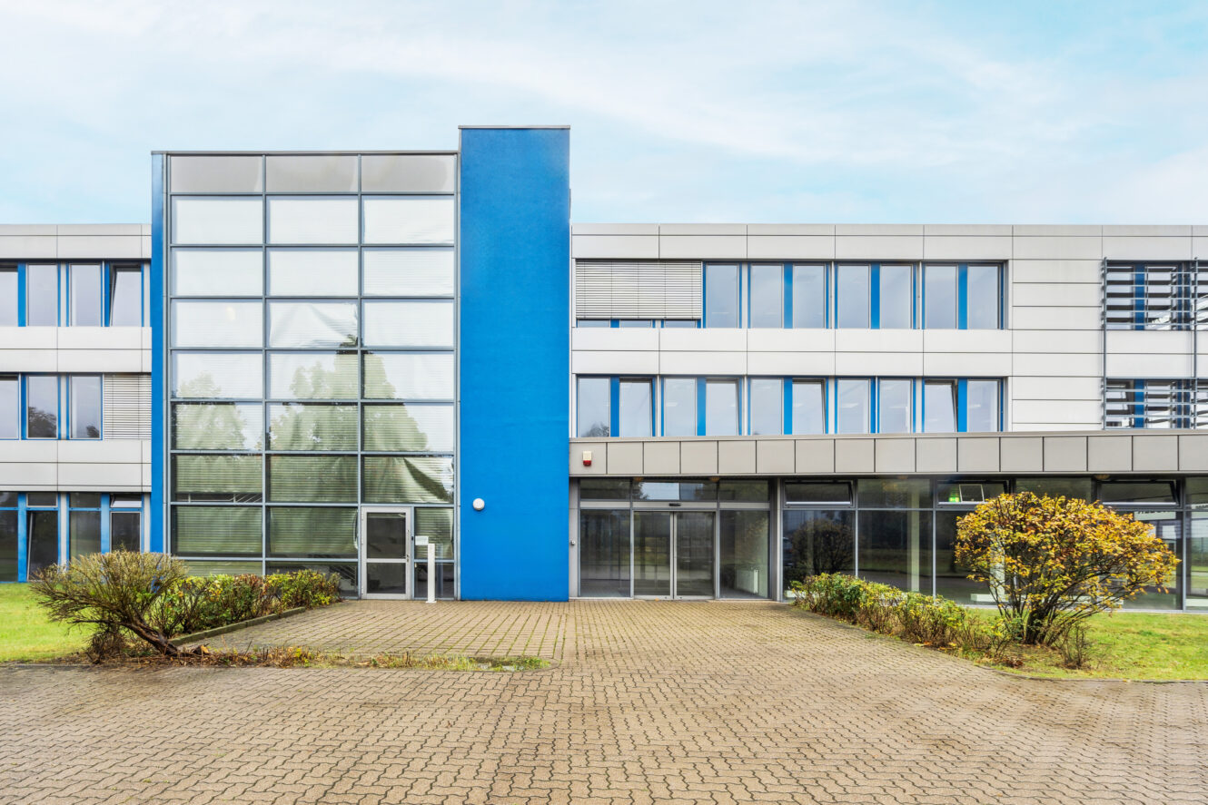 Modernes Geschäfts- oder Bürogebäude mit großen Glasfenstern, blauen vertikalen Akzenten, gepflastertem Innenhof und spärlicher Bepflanzung vor dem Gebäude.