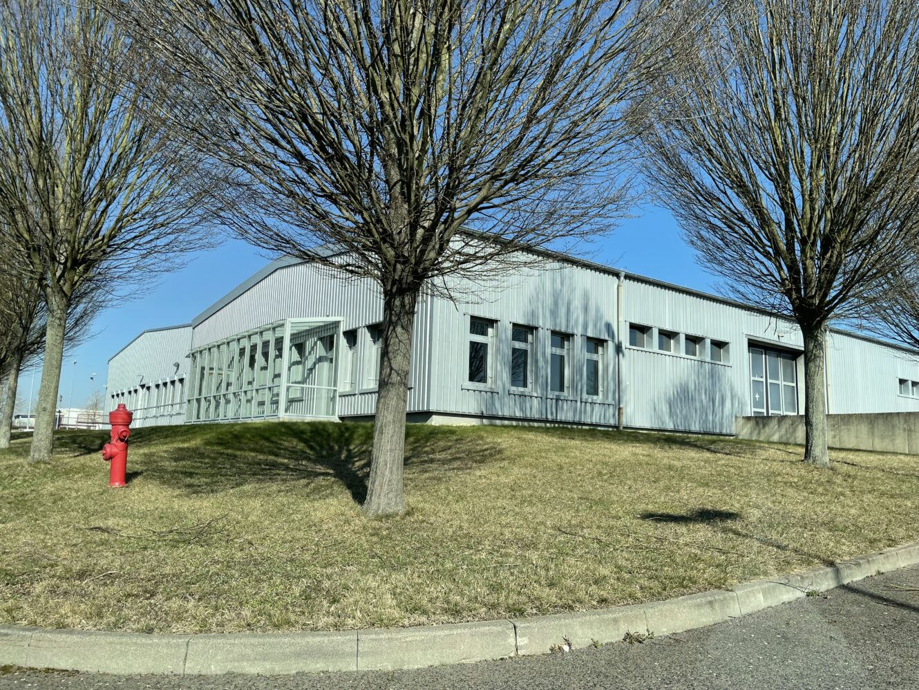 Ein hellgraues Industriegebäude mit großen Fenstern steht an einem grasbewachsenen Hang, flankiert von blattlosen Bäumen und einem roten Hydranten, unter einem klaren blauen Himmel.