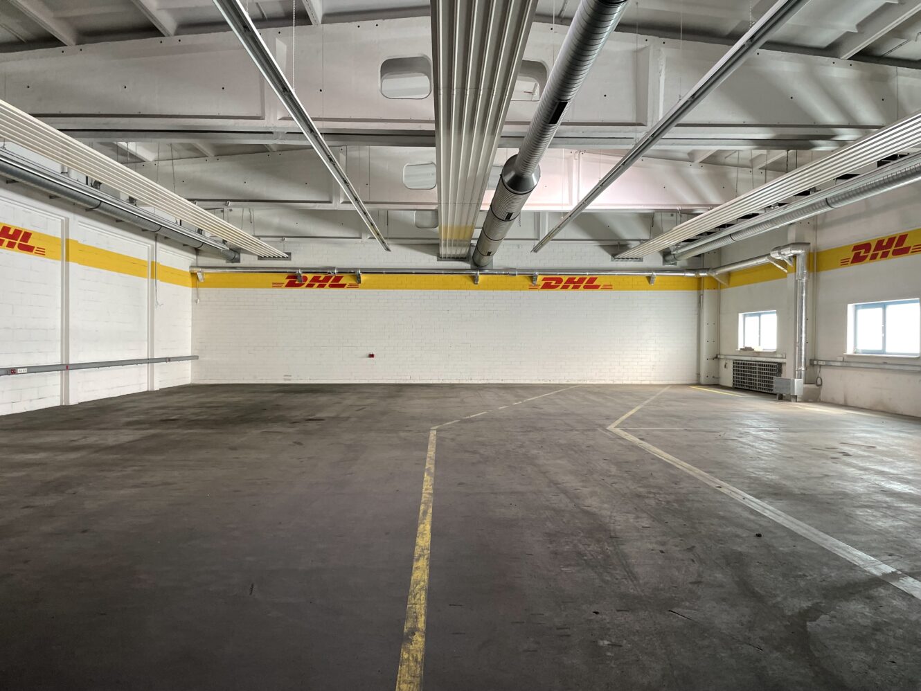 Leeres Lagerhaus mit gelben DHL-Logos an den Wänden, großen Deckenrohren, Betonboden und Fenstern auf der rechten Seite, die Tageslicht hereinlassen.