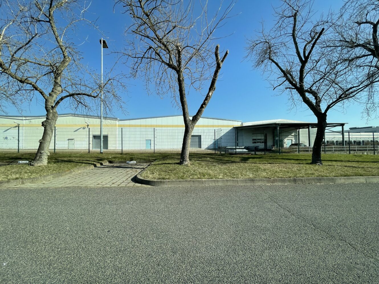 Ein großes Industriegebäude mit einer hellen Fassade, umgeben von blattlosen Bäumen und einer Grasfläche, unter einem klaren blauen Himmel.