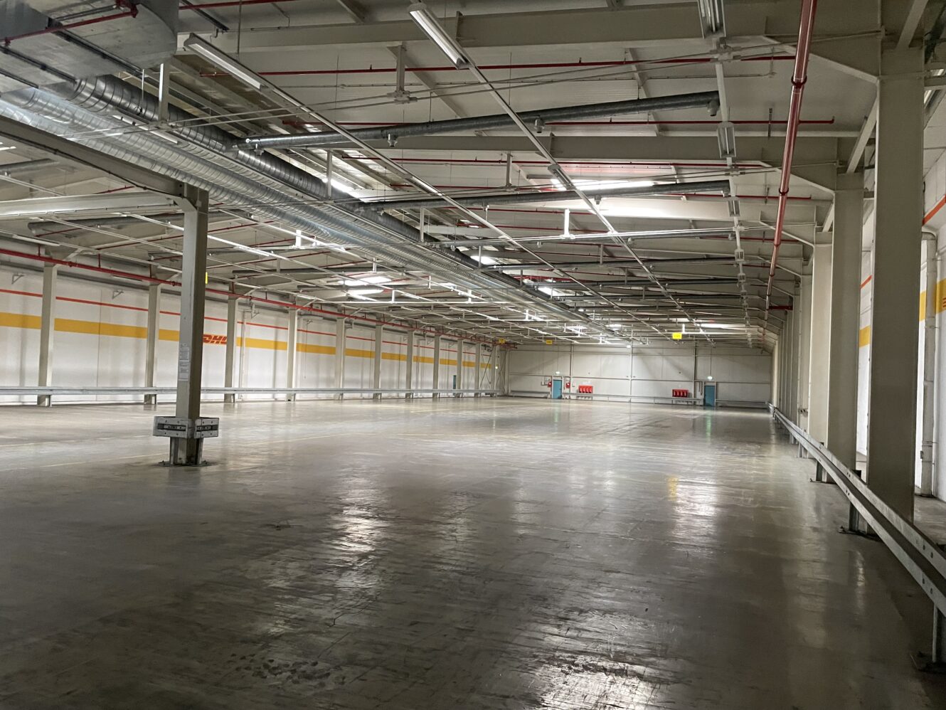 Geräumiges, leeres Industrielager mit Betonböden, hohen Decken, Deckenbeleuchtung und sichtbaren Lüftungsschächten.