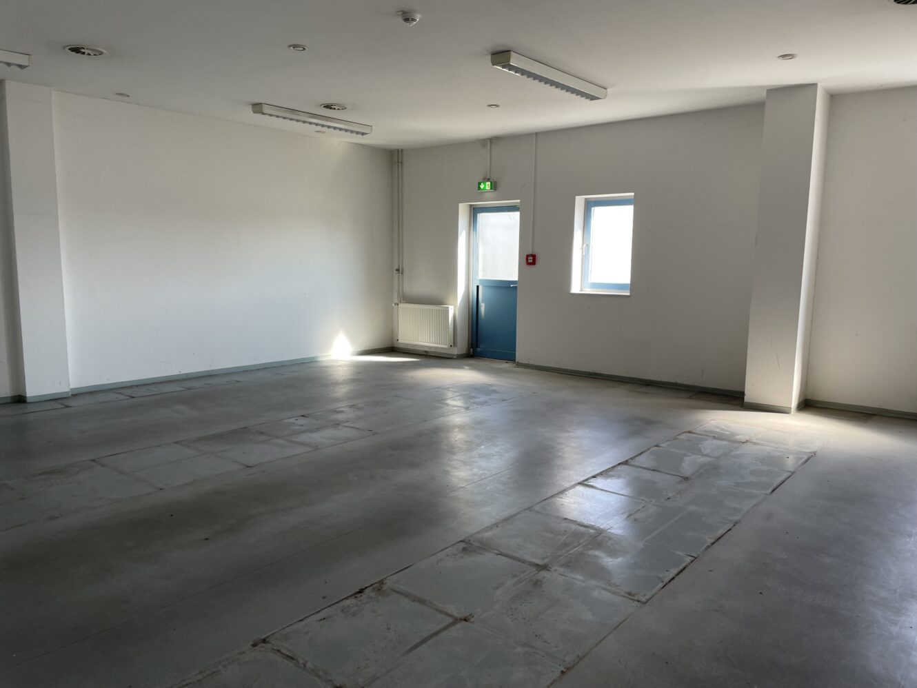 Ein leerer Raum mit weißen Wänden, Betonboden und fluoreszierenden Deckenleuchten. Es gibt eine blaue Tür, ein kleines Fenster und einen Heizkörper an der gegenüberliegenden Wand.