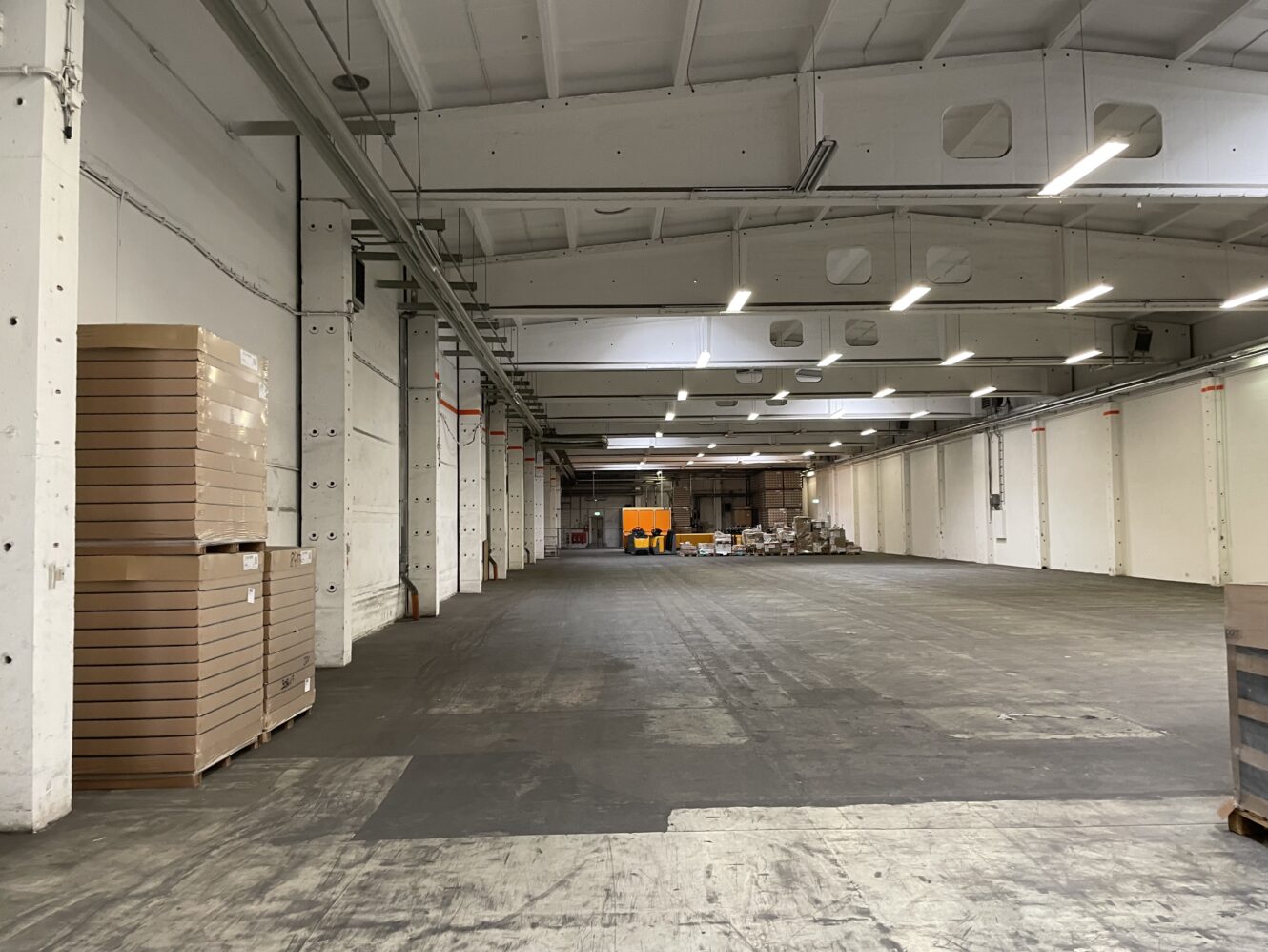 Große, größtenteils leere Lagerhalle mit hohen Decken, gestapelten Kartons auf der linken Seite und verstreuten Materialien und Paletten im hinteren Bereich. Helle Oberlichter erhellen den Raum.