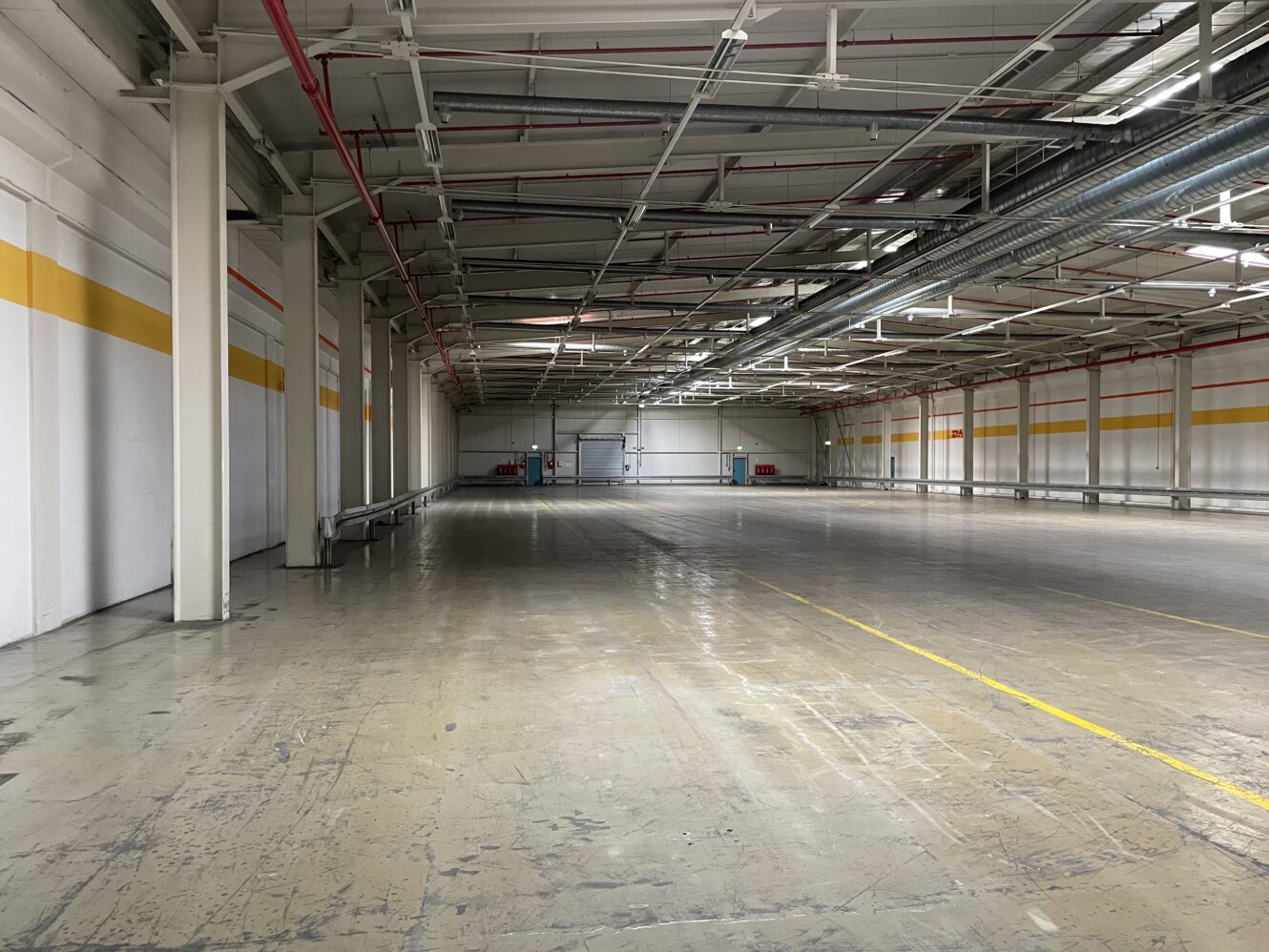 Große, leere Industrielagerhalle mit Betonboden, hoher Decke, freiliegenden Rohren und gelben Linien an den Wänden und auf dem Boden.