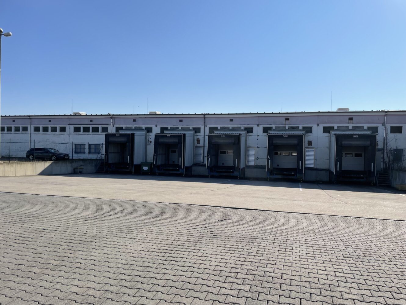 Eine Reihe von fünf Laderampen in einem Lagerhaus, links davon ein geparktes Auto und davor eine leere gepflasterte Fläche, unter einem klaren blauen Himmel.