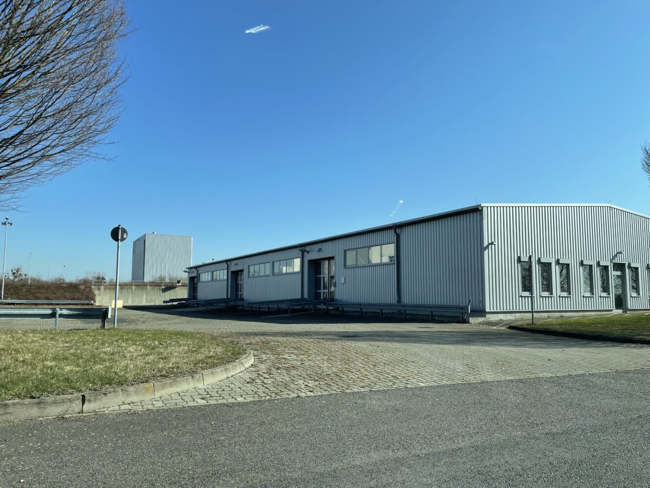 Ein großes, graues Industrielager mit mehreren Fenstern steht neben einer gepflasterten Straße unter einem klaren blauen Himmel. Im Vordergrund befinden sich Gras und ein blattloser Baum.