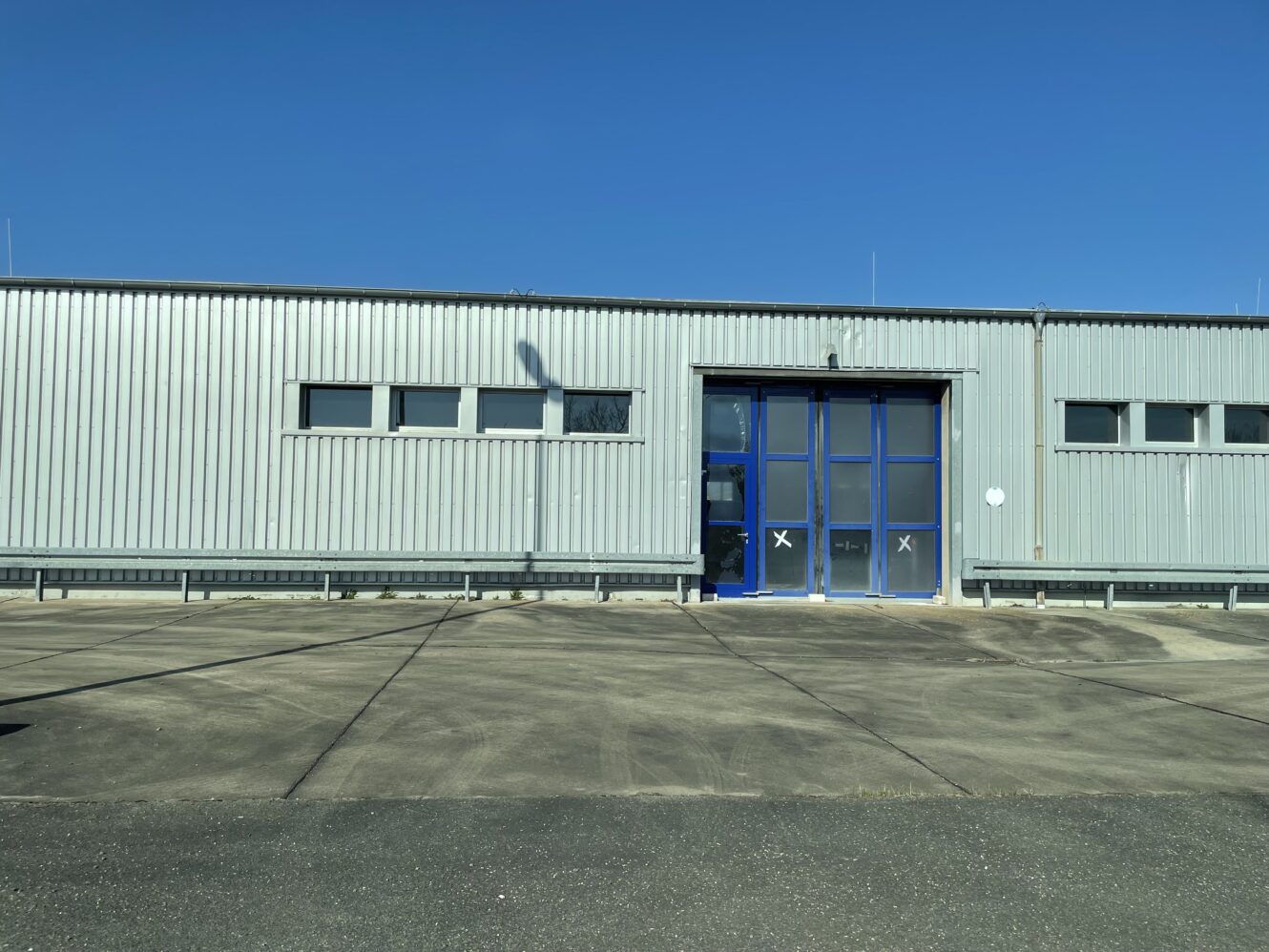 Ein großes Industrielager mit Metallverkleidung, blau umrahmten Fenstern und einer blauen Eingangstür, neben einem leeren Betongrundstück unter einem klaren blauen Himmel.