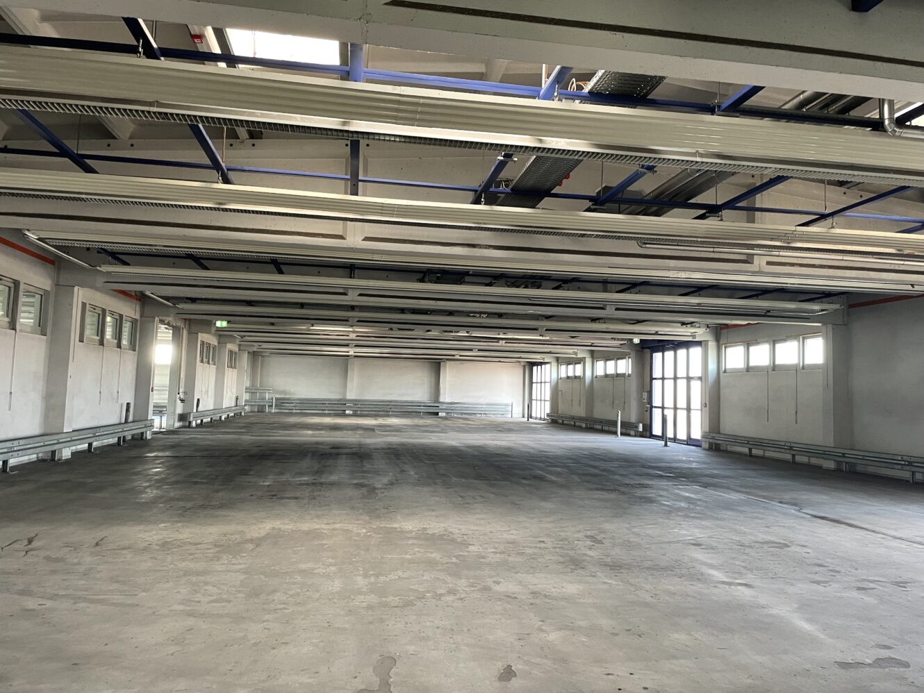 Leeres Industrielager mit Betonboden, sichtbaren Deckenbalken und Fenstern an den Seitenwänden. Durch mehrere Fenster und Türen fällt natürliches Licht ein.