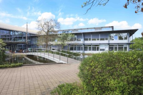 Modernes Bürogebäude mit großen Fenstern und einer Metallbrücke, die über einen Teich führt, umgeben von Grünflächen und einem gepflasterten Gehweg unter einem teilweise bewölkten Himmel.