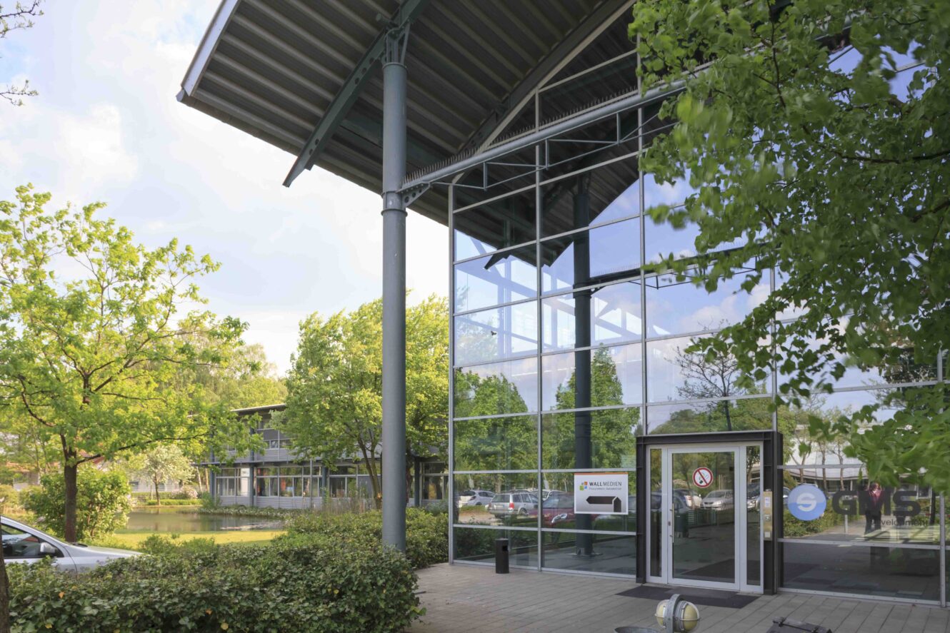 Modernes Glasgebäude mit Metalldach, umgeben von Bäumen und einem Teich; die Autos sind seitlich geparkt, und in der Nähe des Eingangs sind Schilder zu sehen.