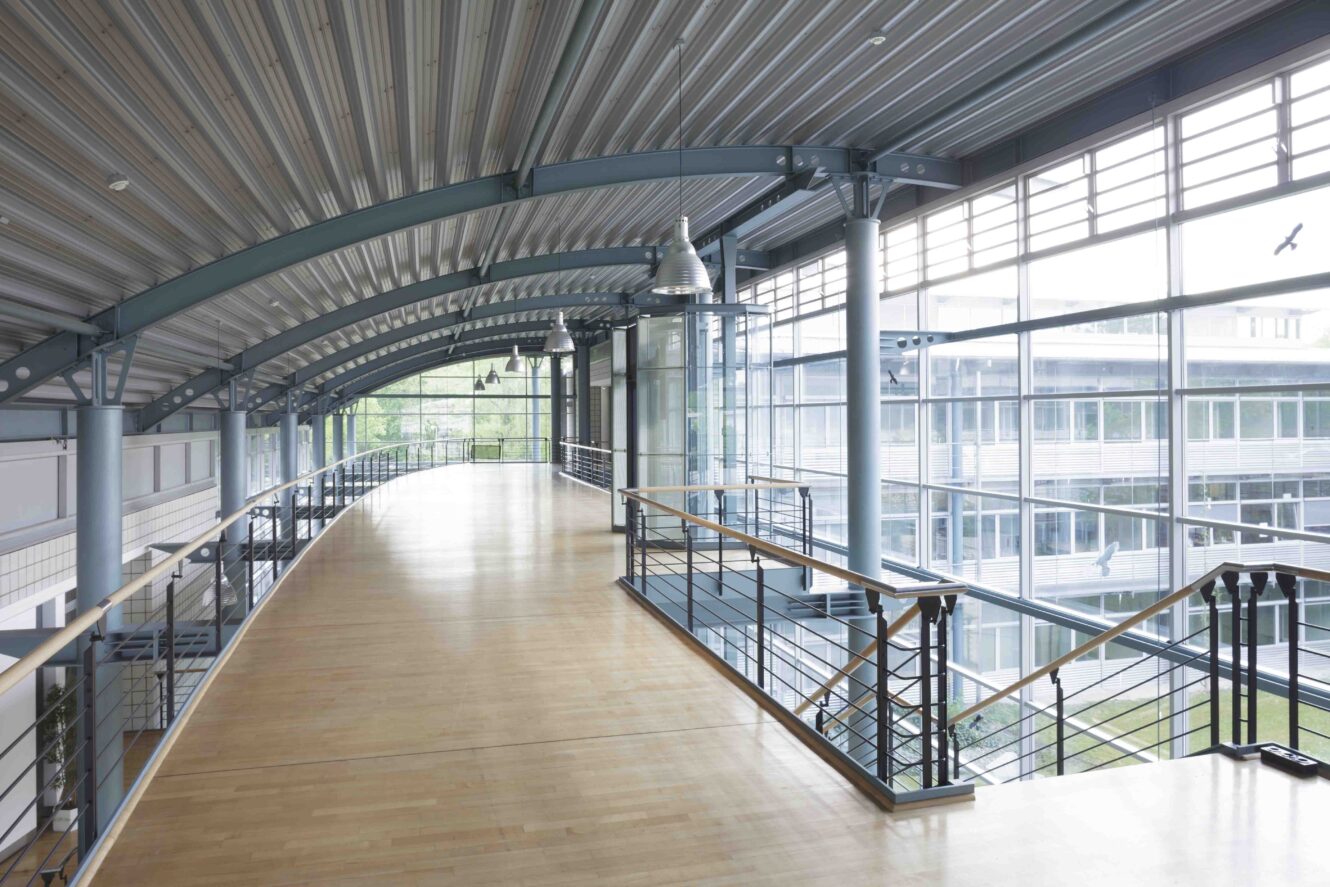 Geräumiger Innengang mit Holzböden, Metallgeländern, großen Fenstern und gewölbten Deckenbalken mit Blick auf einen modernen Innenhof.