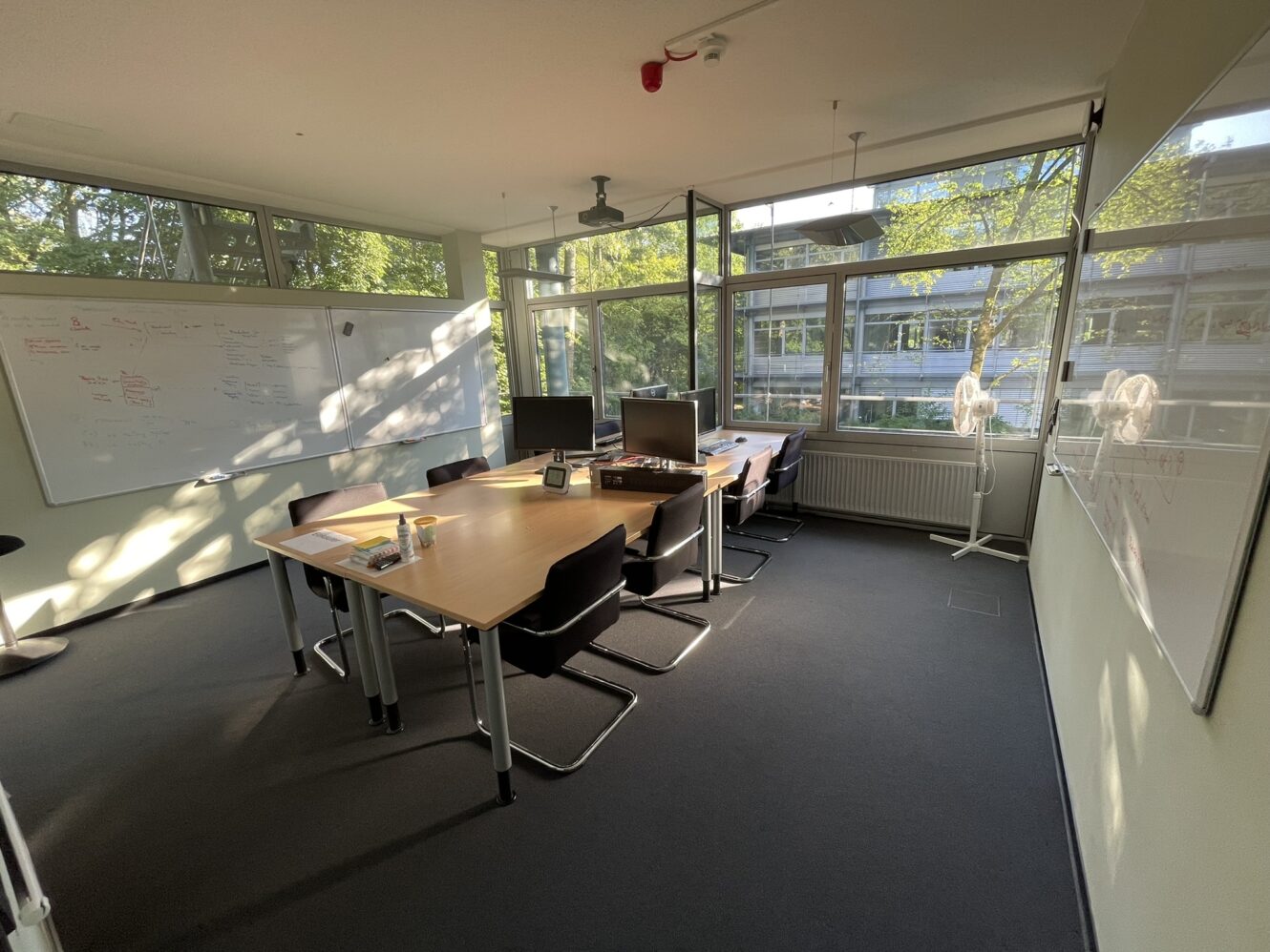 Ein modernes Büro mit großen Fenstern, einem Konferenztisch mit sechs Stühlen, zwei Monitoren, zwei Ventilatoren und Whiteboards an den Wänden. Sonnenlicht strömt in den Raum.