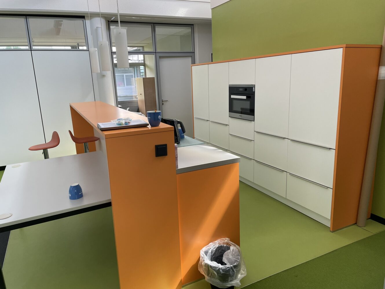 Moderne Büroküche mit orangefarbenen und weißen Schränken, einem eingebauten Ofen, einer Theke mit Hockern und einem Tisch mit blauen Bechern. Der Mülleimer steht neben dem Tresen.