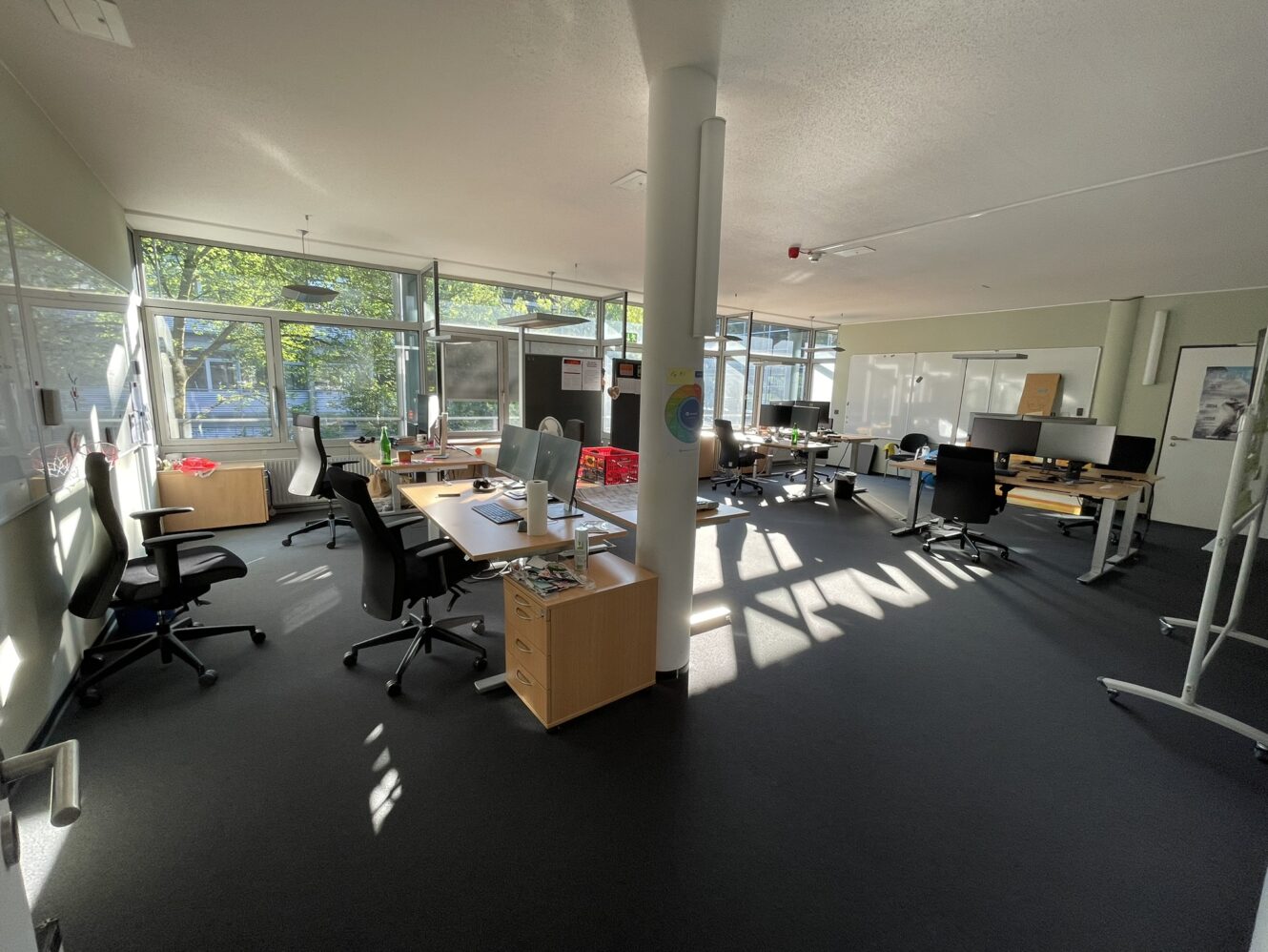 Großraumbüro mit mehreren Schreibtischen, Bürostühlen, Computern und großen Fenstern, die natürliches Licht hereinlassen. Das Büro wirkt aufgeräumt und unbesetzt.