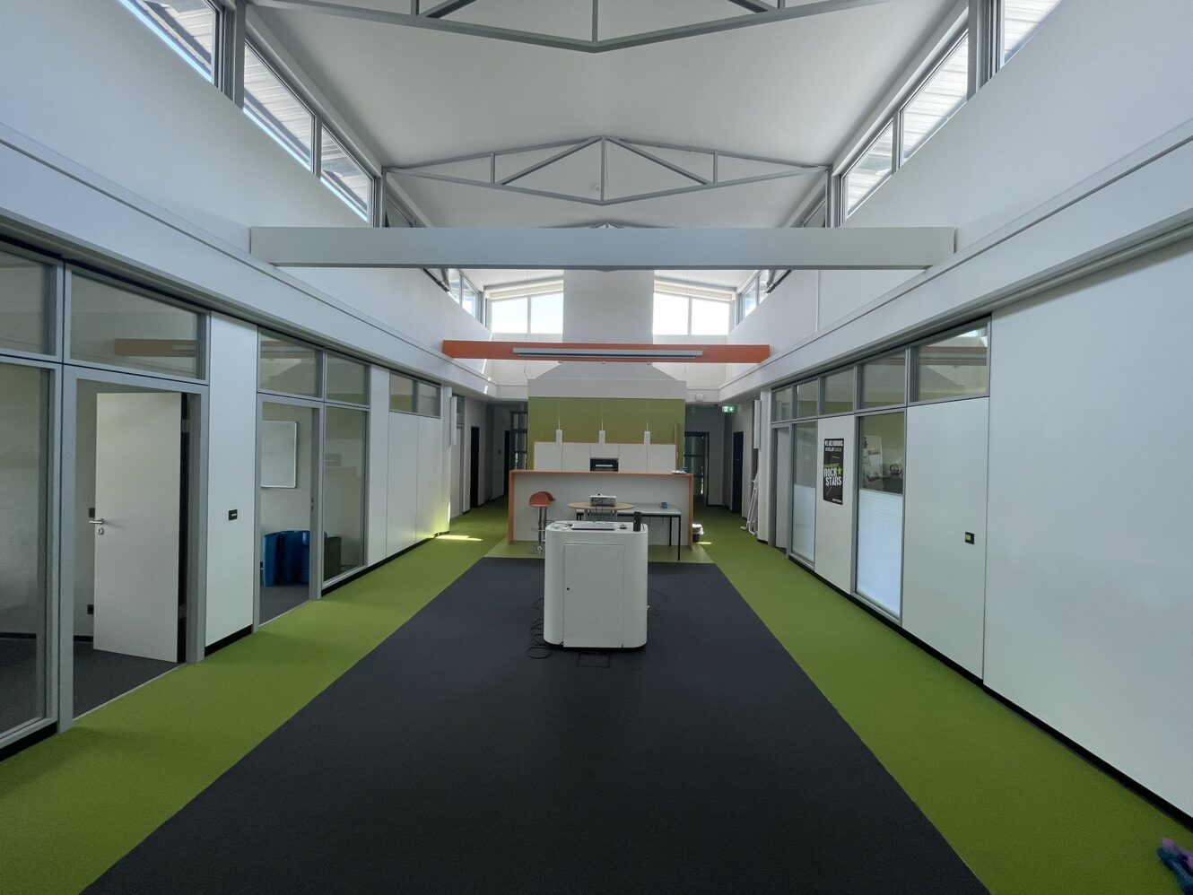 Moderner Büroflur mit grünem und schwarzem Bodenbelag, beidseitig verglasten Räumen und einer zentralen Druckerstation unter hohen Decken mit Sichtbalken.